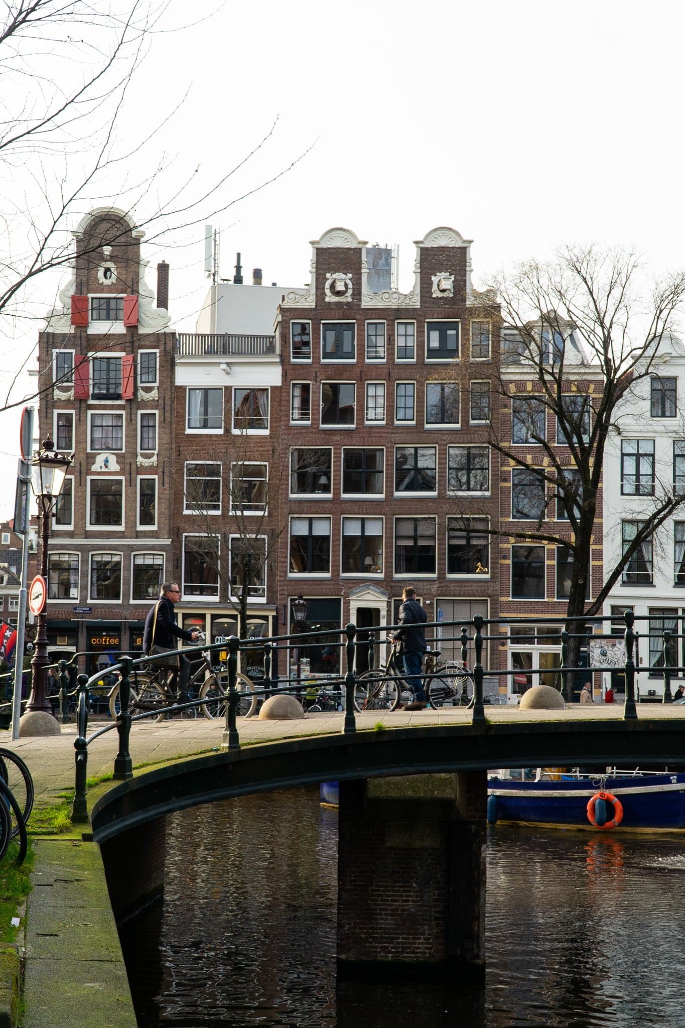 Ornate canal-house gables rising beyond an arched bridge where cyclists cross an Amsterdam canal
