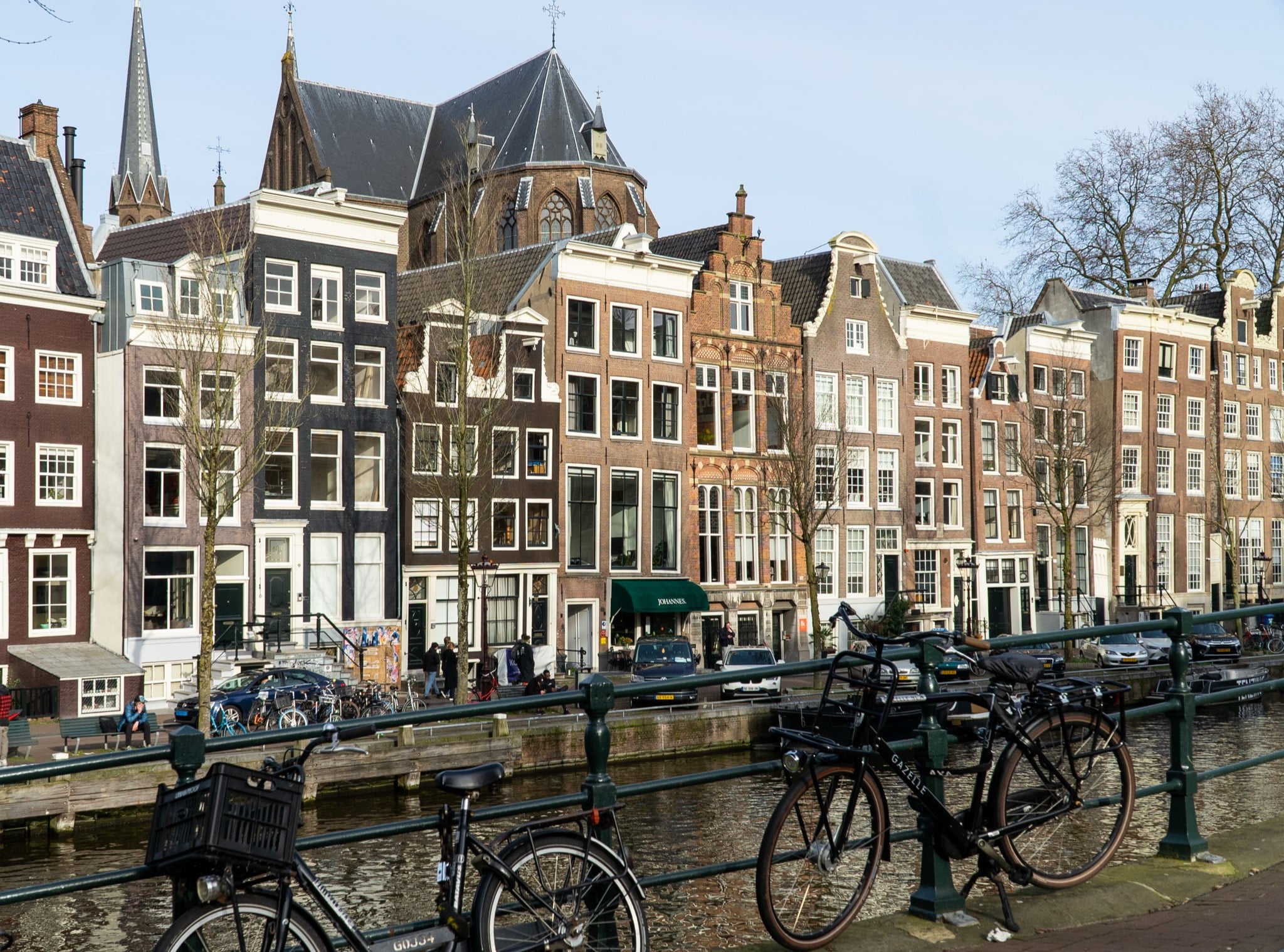 A row of canal houses with a church tower rising behind a bicycle-lined bridge