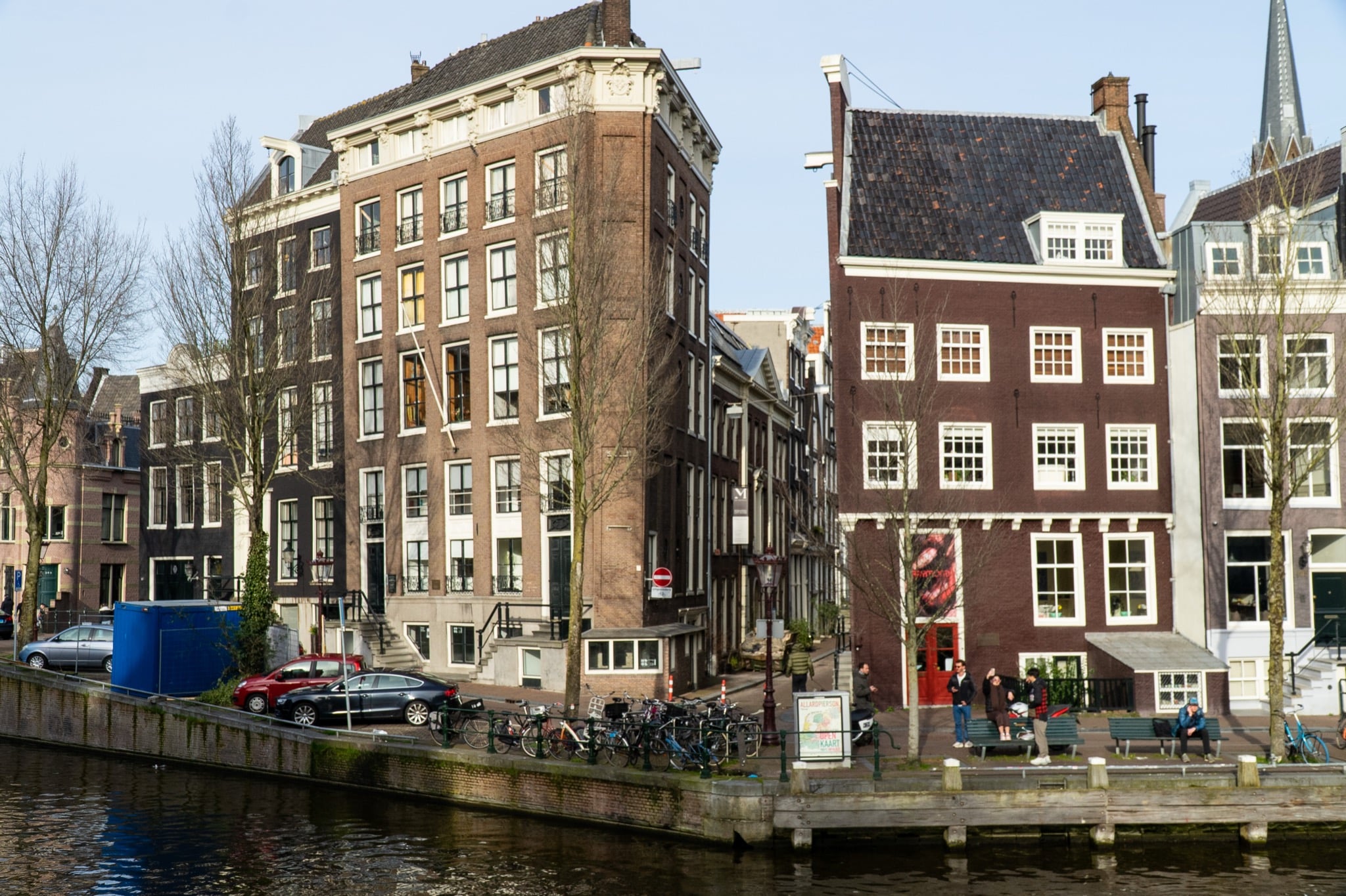 Tall canal houses along a brick quay with parked bicycles and people resting on benches