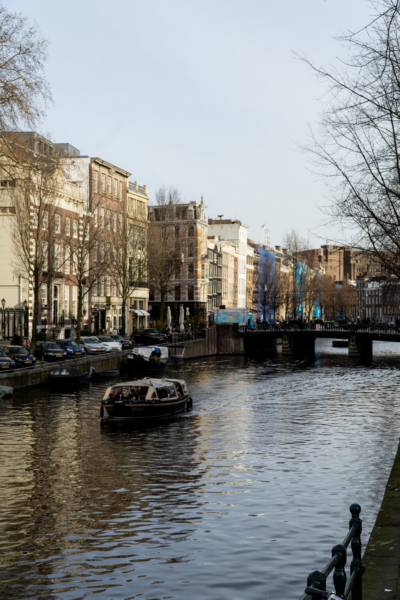 A small tour boat gliding along a canal past a row of sunlit houses toward a bridge