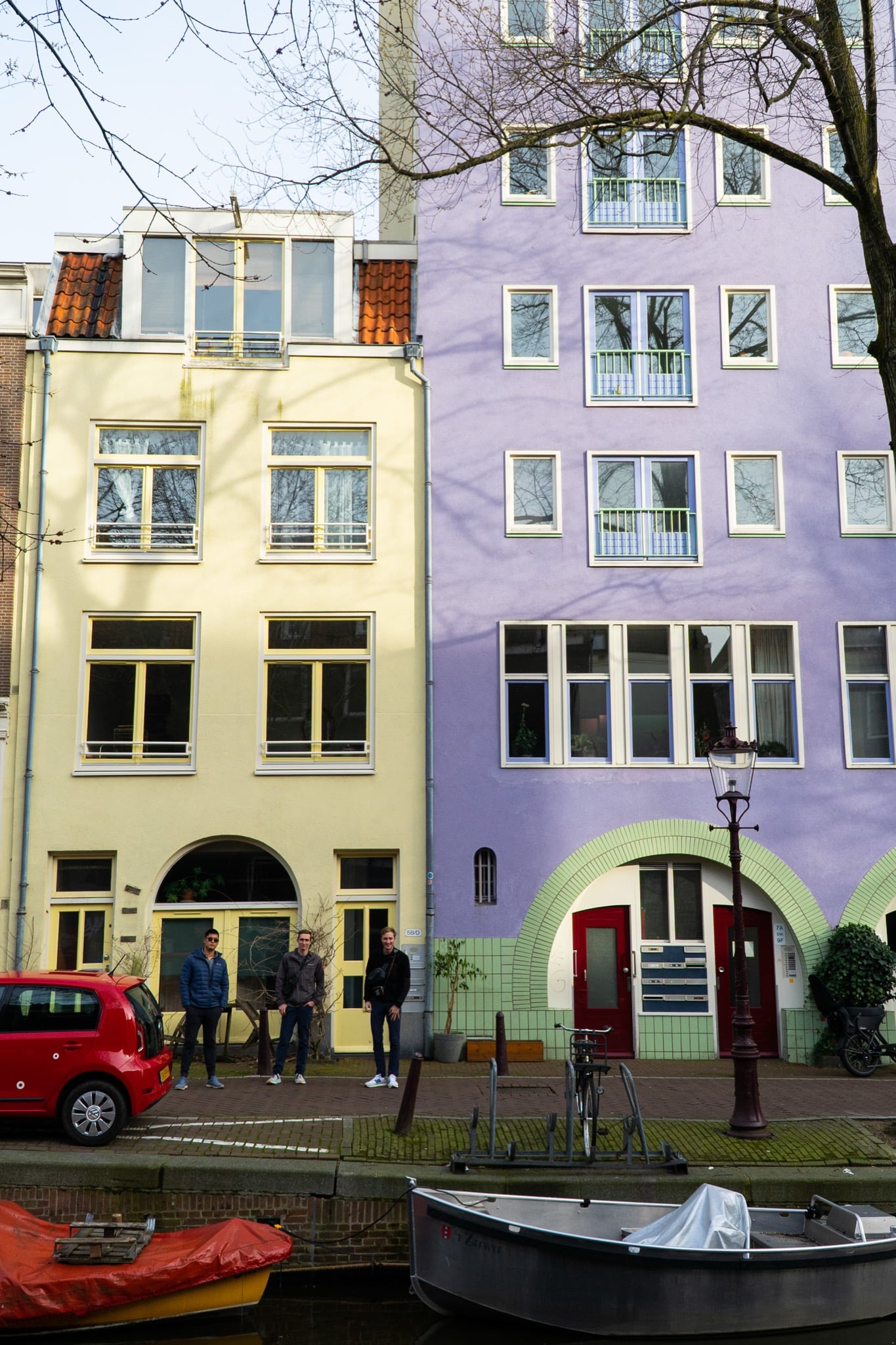 A pale-yellow and lilac canal house with green-arched doorways, three friends posing on the quay