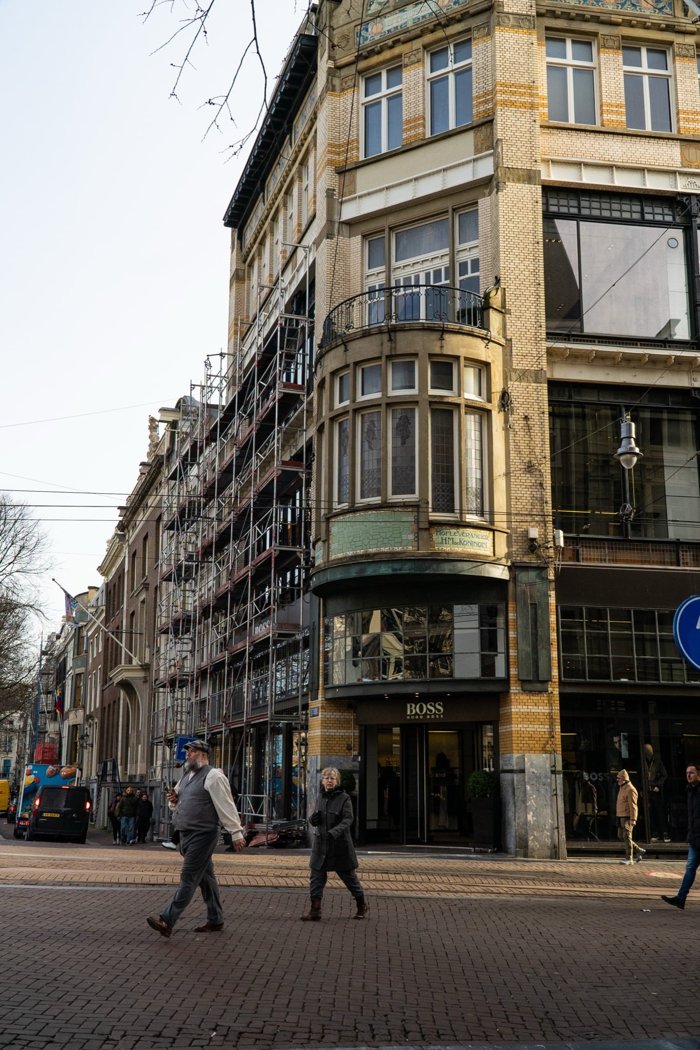 A tiled Art Nouveau corner building under scaffolding, pedestrians crossing the brick plaza below