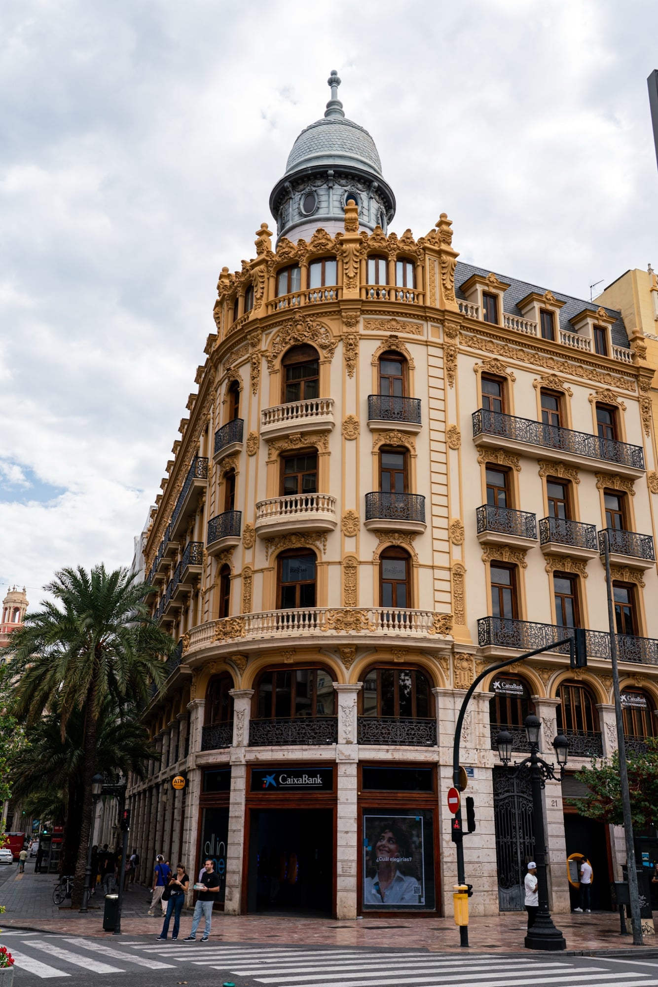 An ornate golden-ochre corner building crowned with a grey dome, palms and a pedestrian crossing below