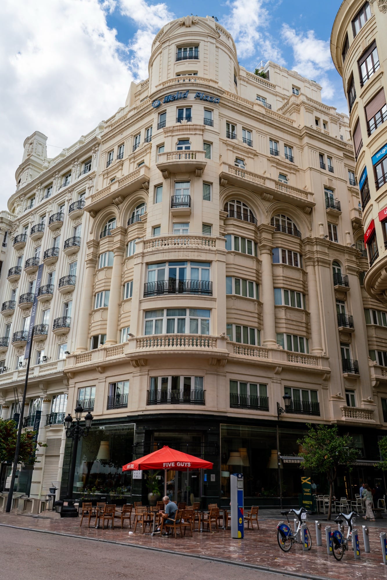 A towering cream apartment building with curved bay windows above a café terrace and its red umbrella