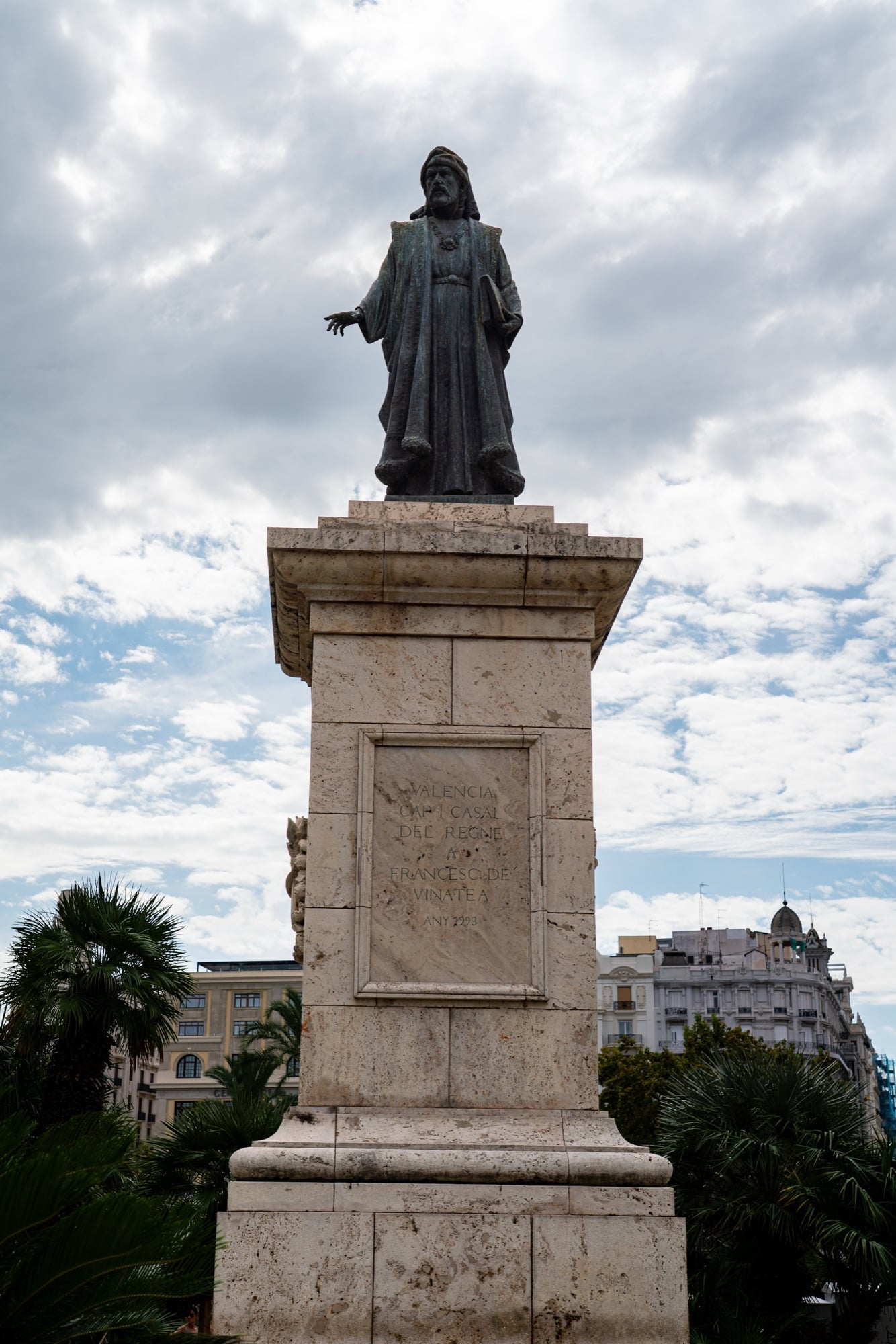 A bronze statue in a long robe atop a tall stone pedestal, palms and rooftops beneath a cloudy sky