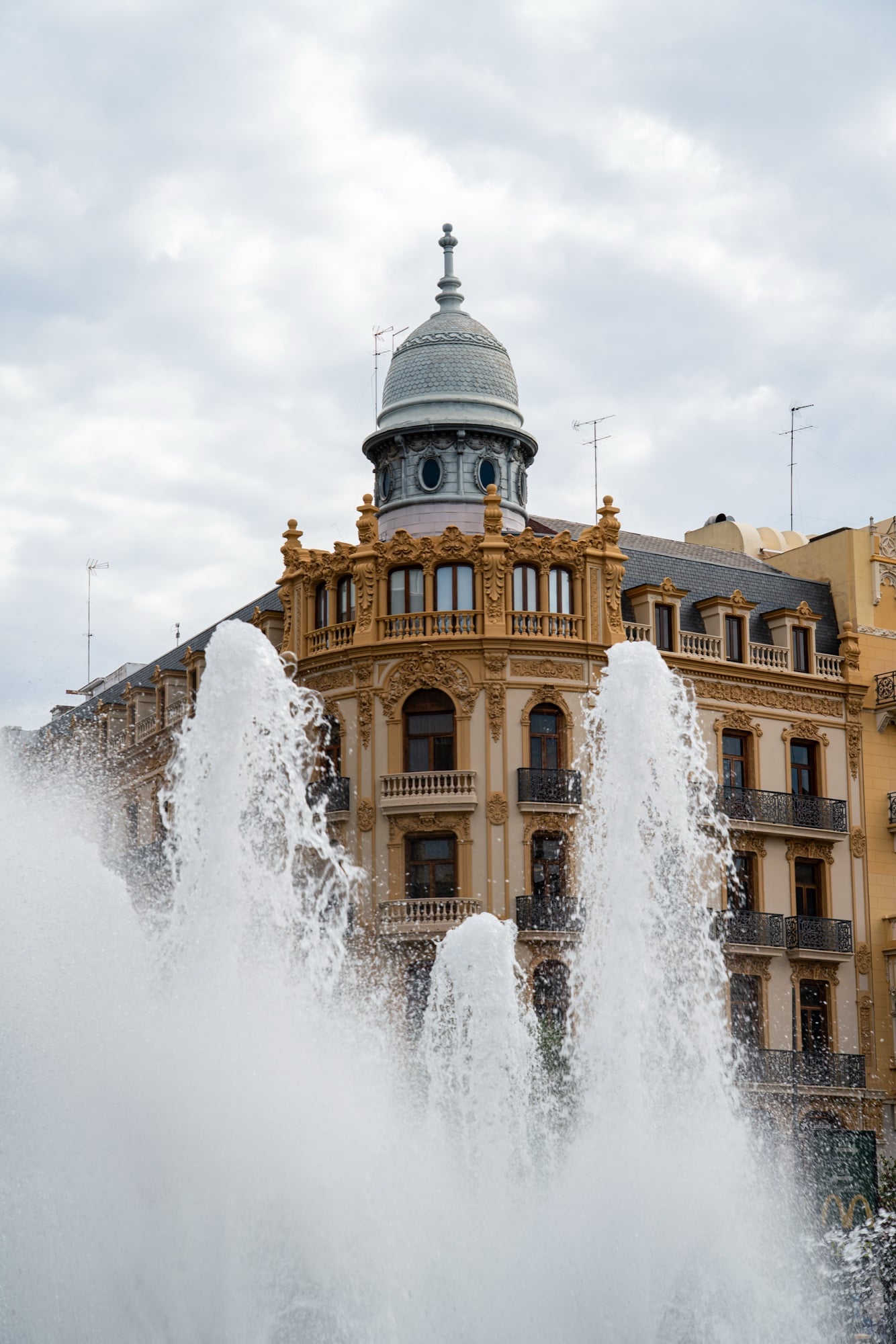 Fountain jets bursting white in front of a golden corner building with a grey dome