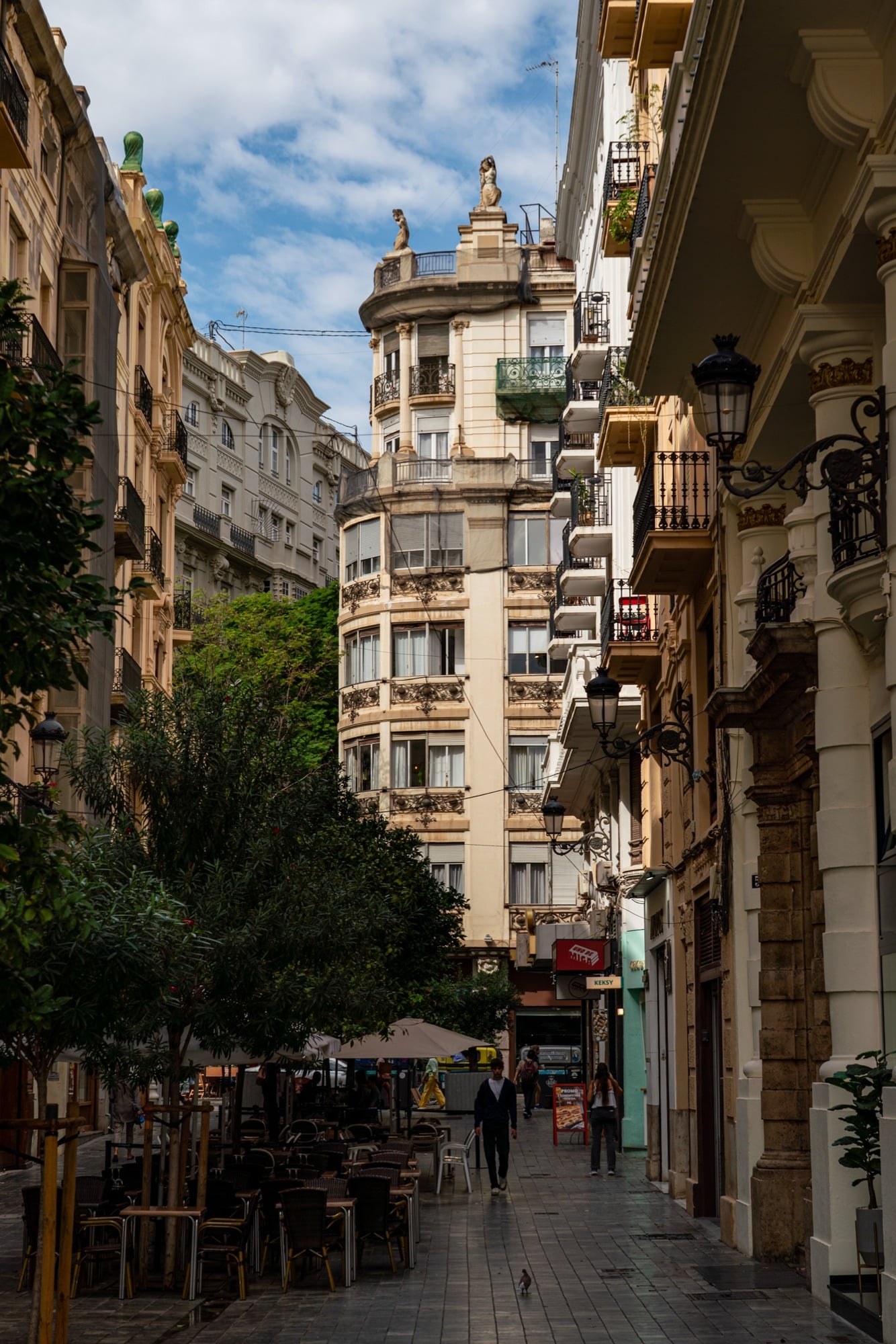 A narrow café lane shaded by trees, a cream round-cornered tower building closing the view