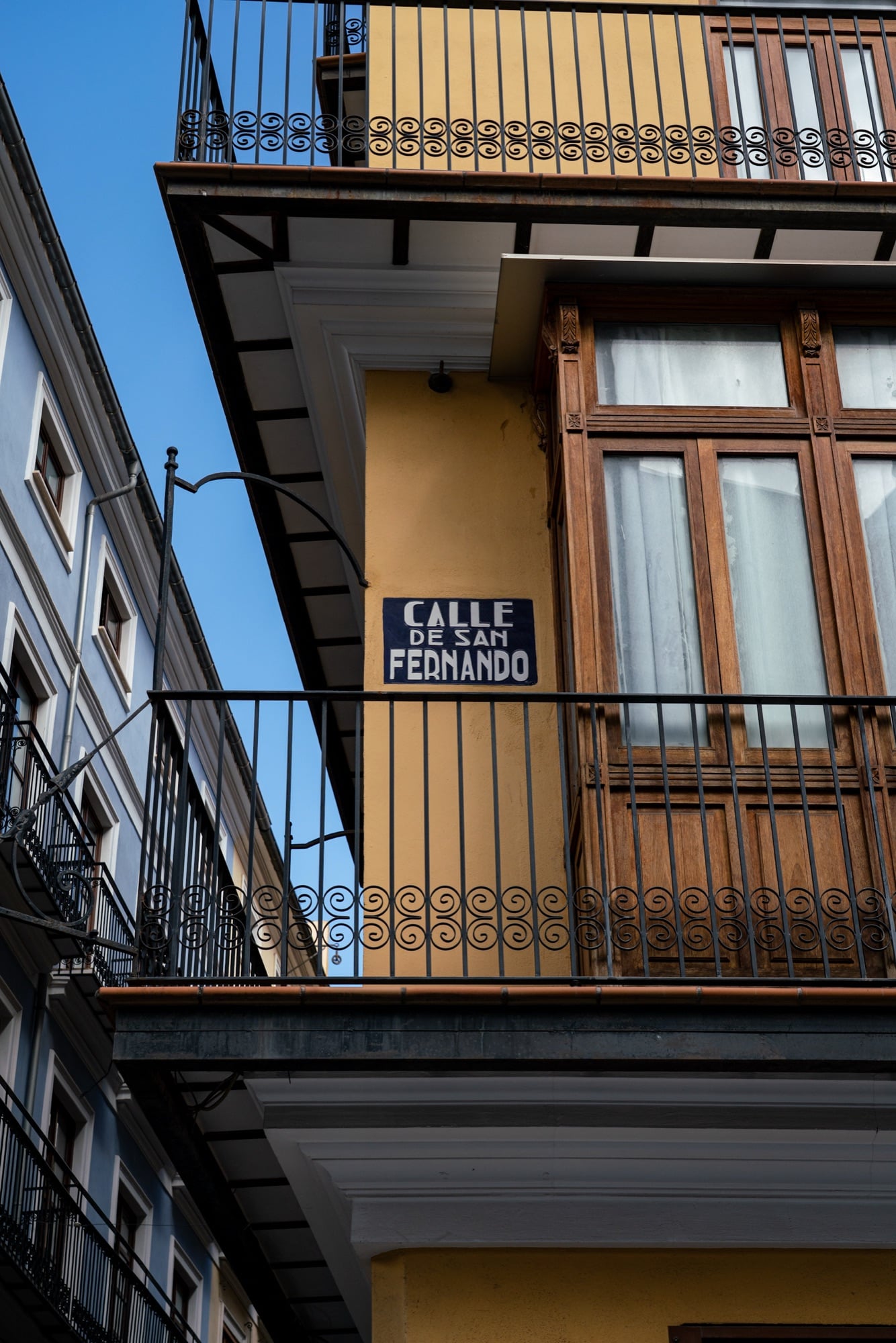 A blue-and-white Calle de San Fernando street sign on a yellow building beneath a wooden enclosed balcony