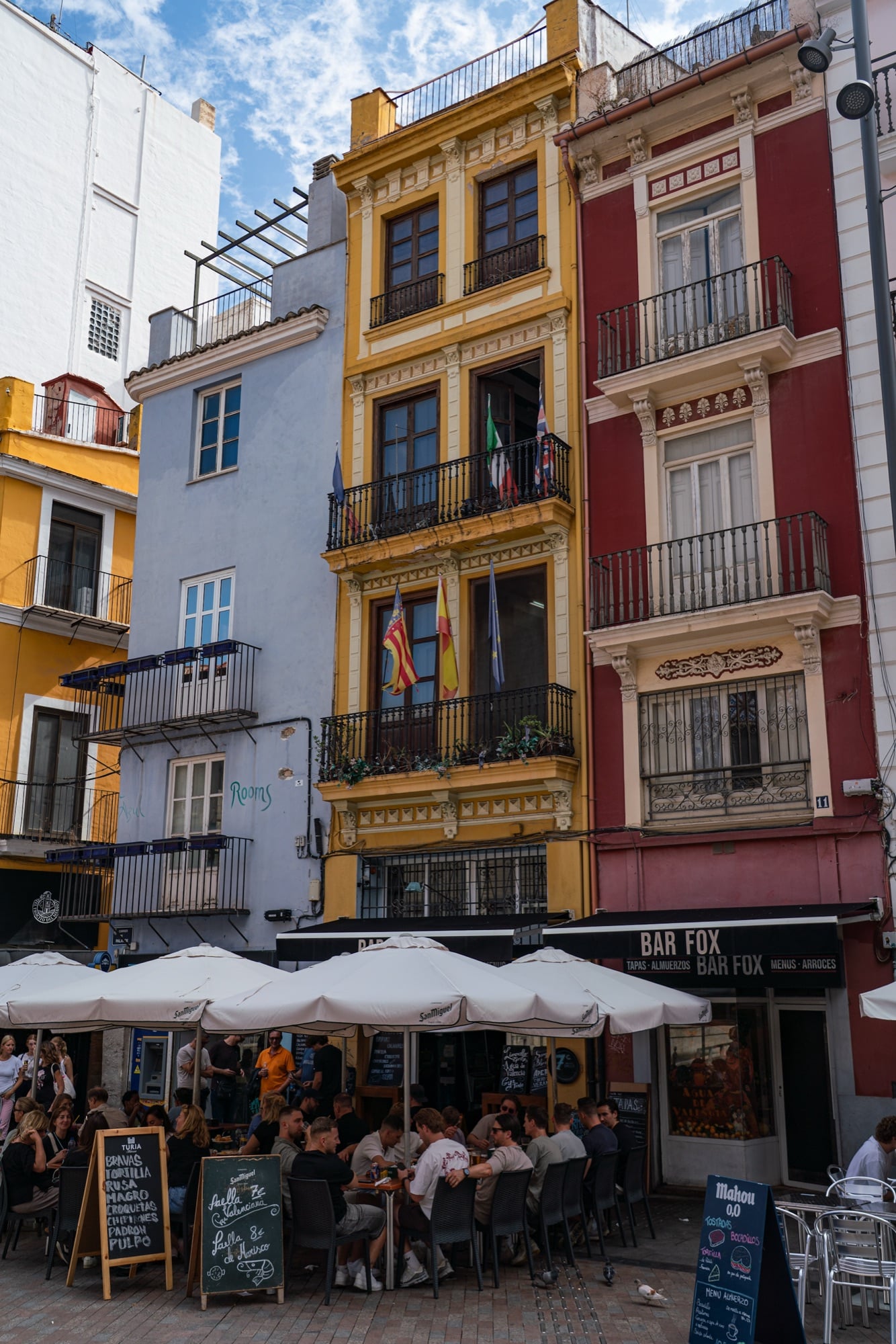 Yellow, red and white townhouses with iron balconies above a busy bar terrace and its umbrellas
