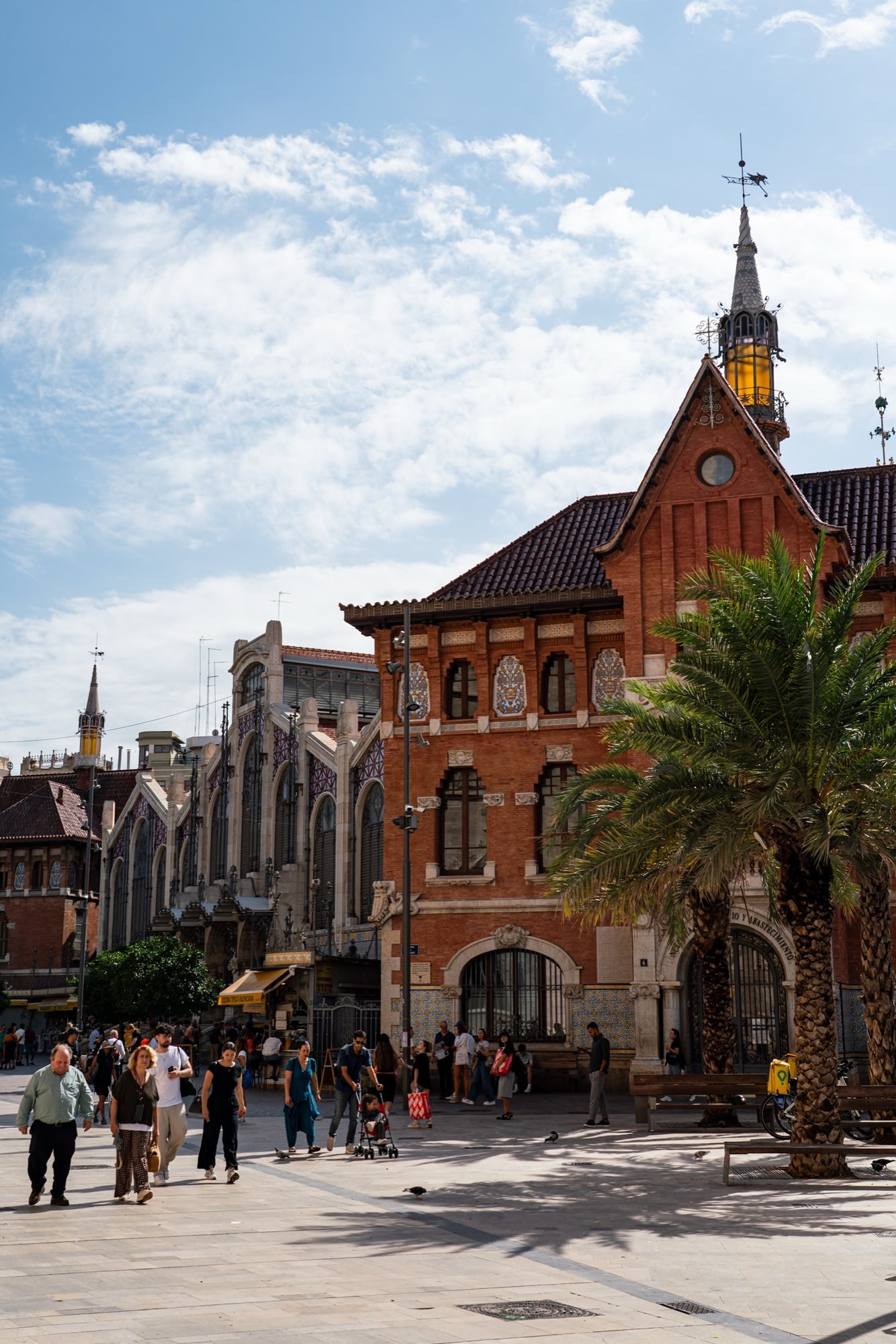 A red-brick building with a spired turret behind a palm tree, people crossing the sunlit square in front