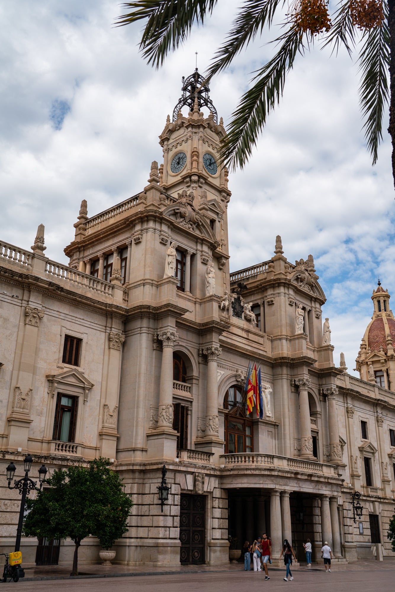 The clock tower of Valencia City Hall framed by drooping palm fronds against a cloud-streaked sky