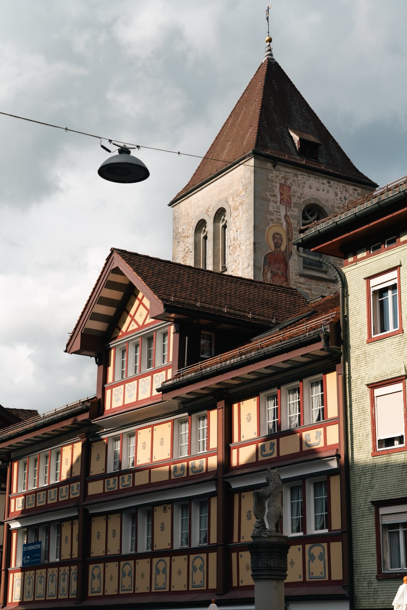 Ornately painted half-timbered facades below a stone church tower bearing a painted saint, a bear-crest fountain column in front