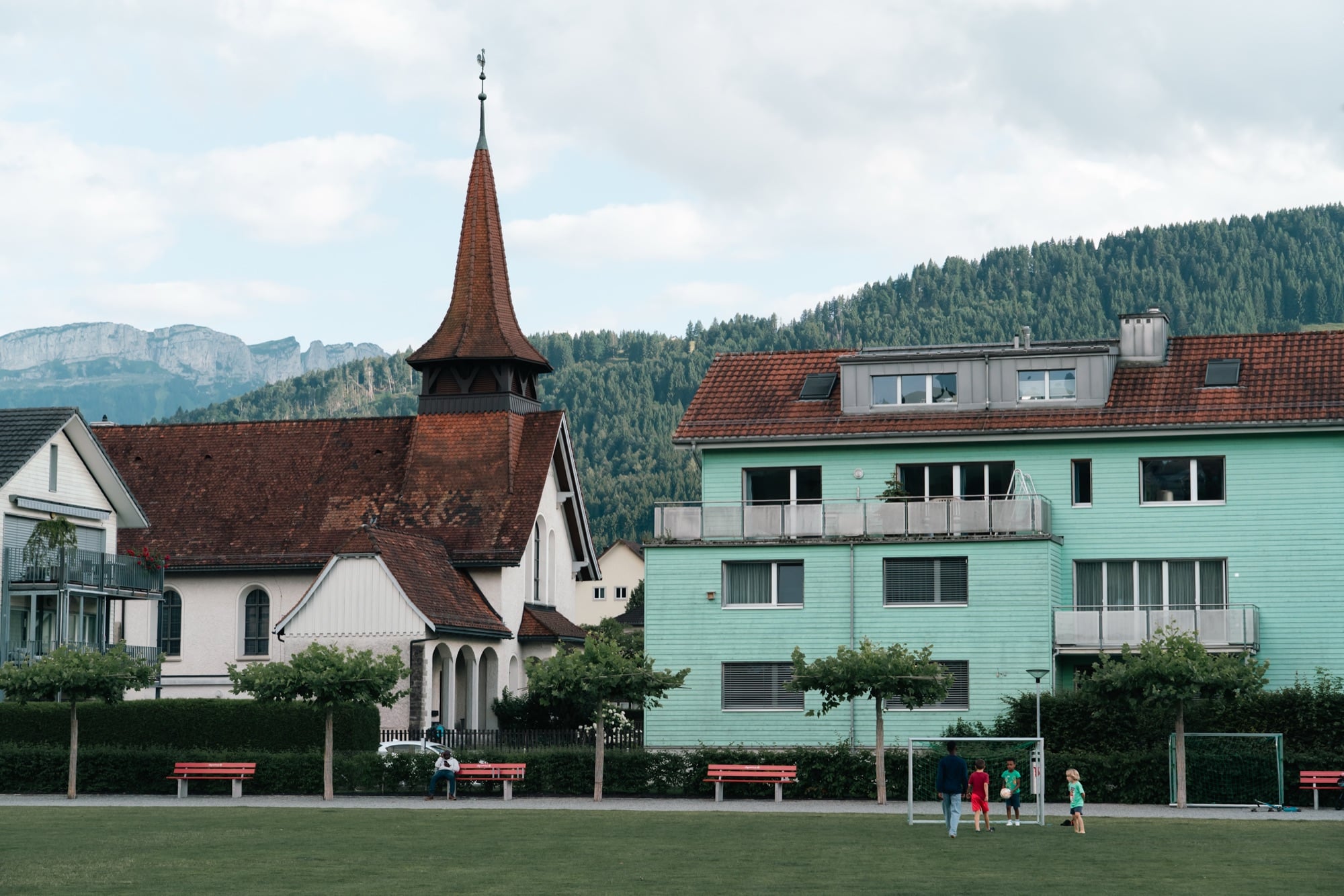 Children playing football on a green pitch below a pointed-spire village church and a mint-green apartment block, a wooded ridge behind