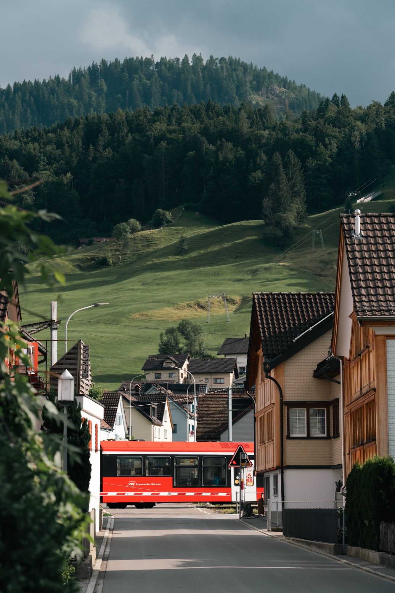 A red Appenzeller Bahnen train passing a level crossing on a village street, a green hillside and dark forest rising behind