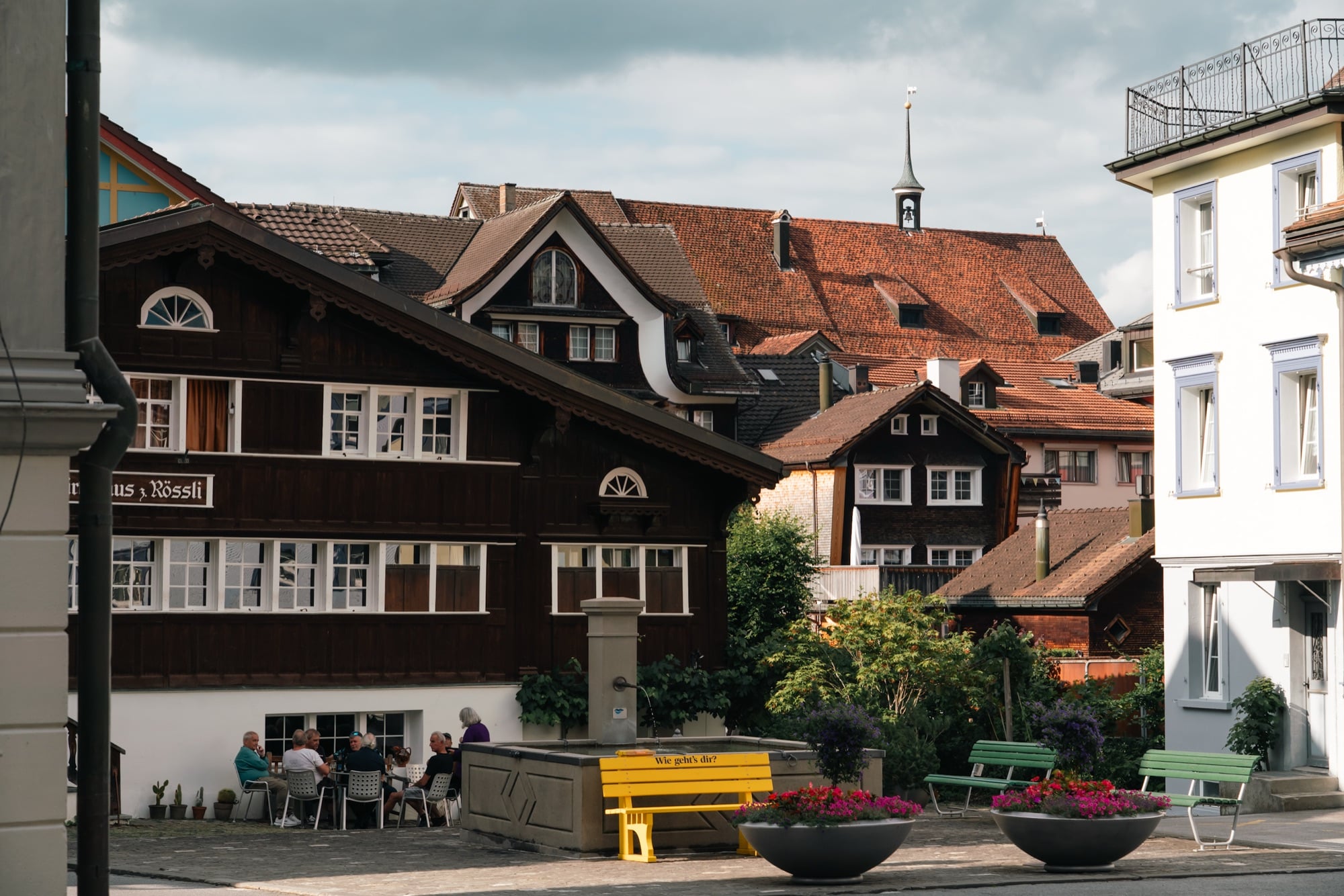 A village square with a stone fountain, a yellow bench reading 'Wie geht's dir?' and flower planters, people at café tables by the wooden Gasthaus zum Rössli