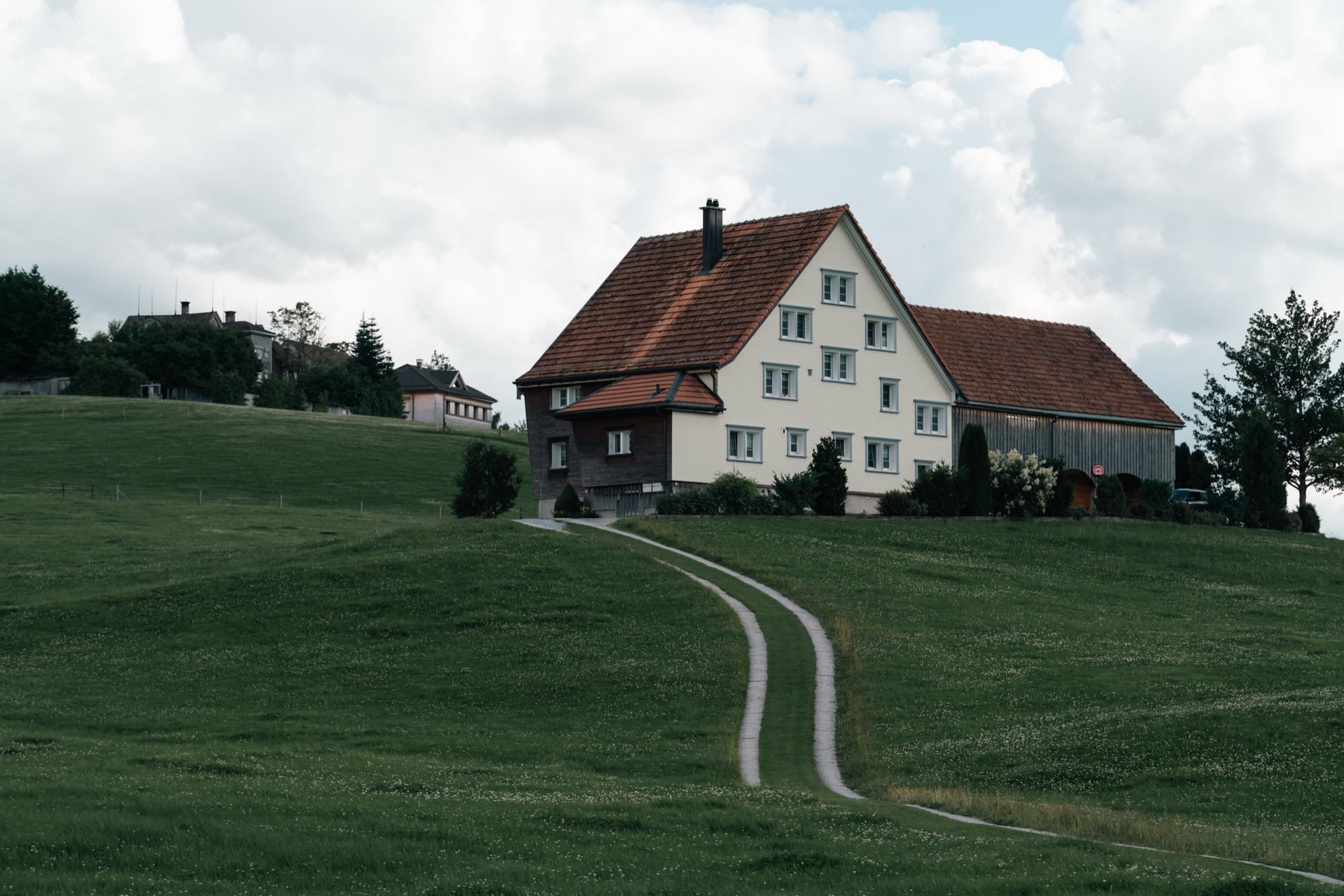 A cream Appenzell farmhouse with a steep red-tiled roof on a green rise, a twin-track path curving up through the meadow to it