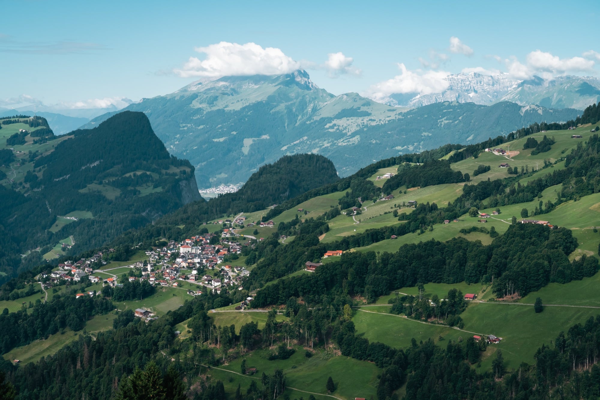 A wide alpine panorama over the Prättigau, villages and farmhouses scattered across green slopes below layered mountains and a snow-streaked summit