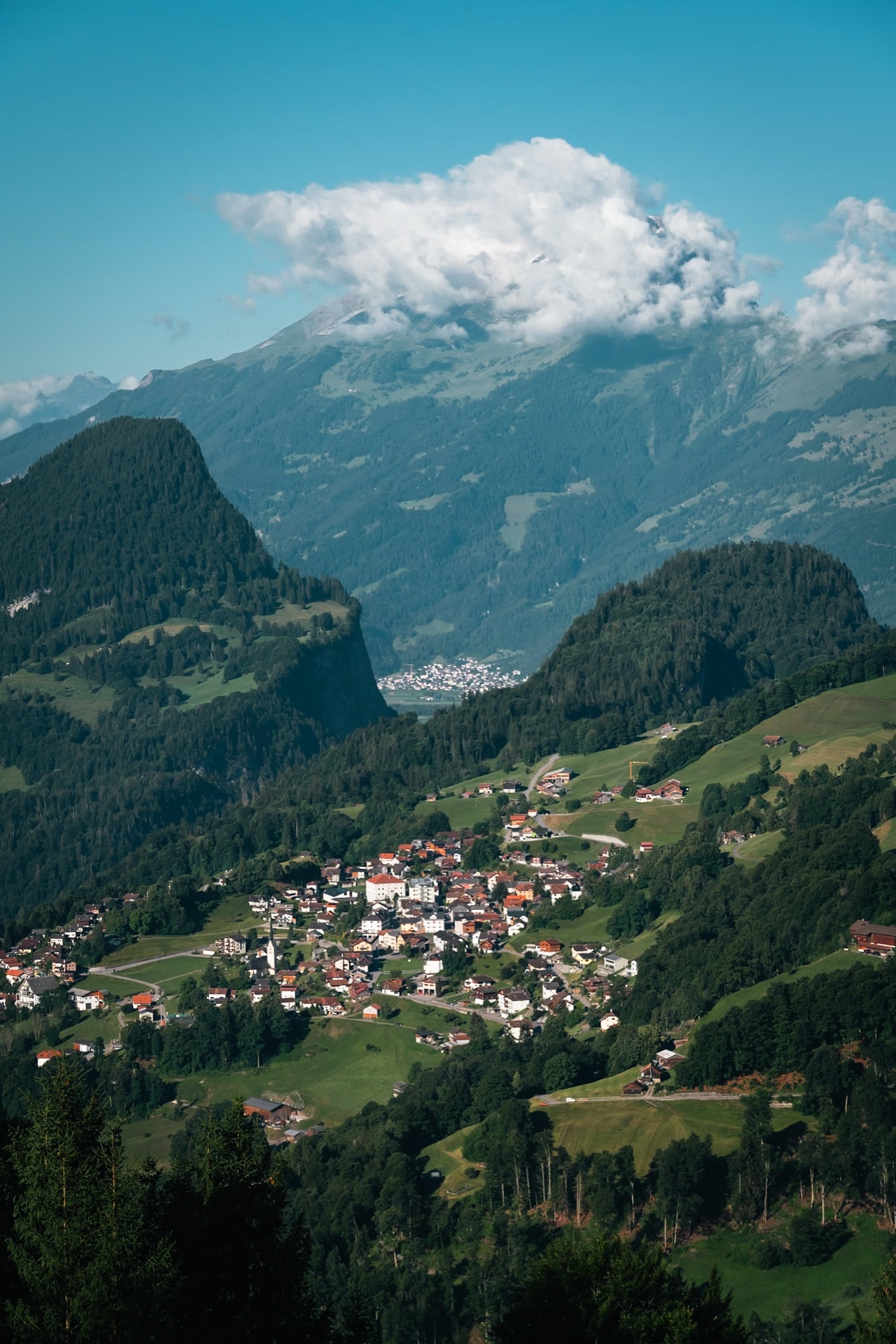 A steep view down into the Prättigau valley, the village of Fanas on green slopes between forested crags below a cloud-capped peak